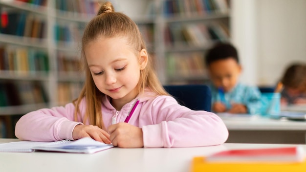 Nella classe della scuola elementare ritratto di una studentessa intelligente che scrive su un taccuino seduto alla scrivania dentro