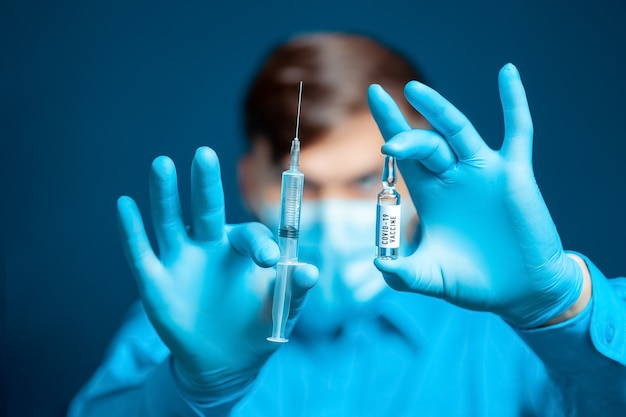 Nelle mani di un medico, un medico con maschera medica e guanti, vestito con un'uniforme blu, una siringa per iniezione e un vaccino contro il coronavirus