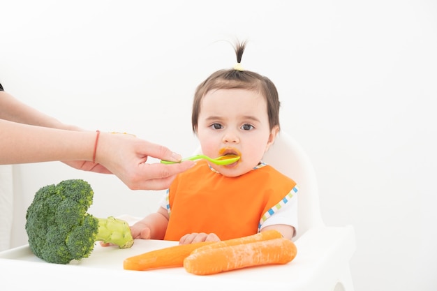 Neonata che si siede in una sedia di Childs che mangia la purea di verdure. La mamma nutre il bambino.