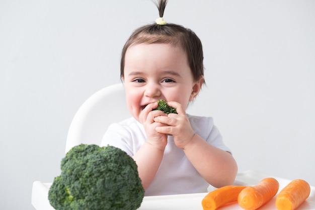 Neonata che si siede nella sedia del bambino che mangia carote e broccoli.