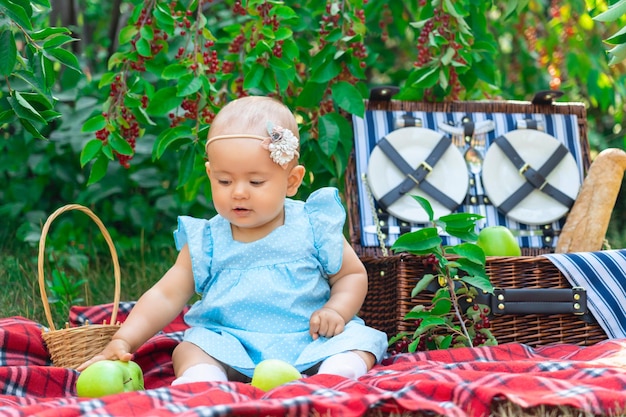 Neonata in un vestito blu su un picnic. Un bambino di 9-12 mesi afferra una mela verde sdraiata su una coperta