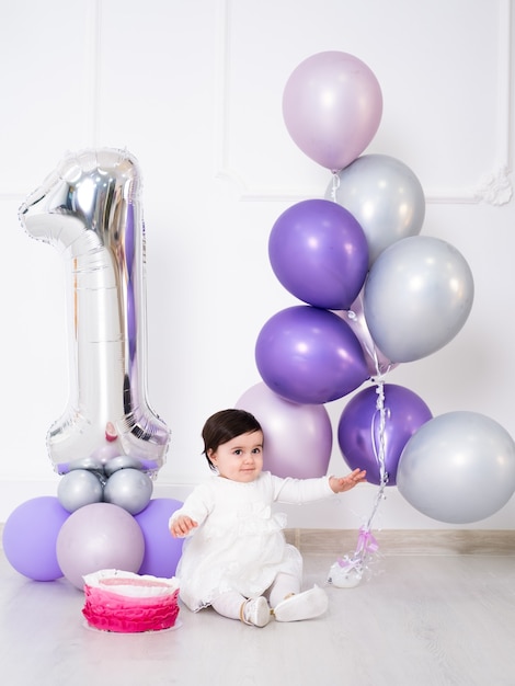 Neonata in vestito bianco che si siede sul pavimento che celebra il suo primo compleanno con torta e palloncini.