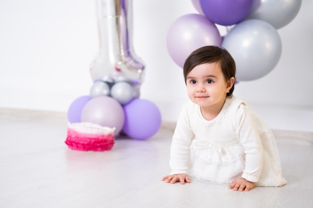 Neonata in vestito bianco che si siede sul pavimento che celebra il suo primo compleanno con torta e palloncini.