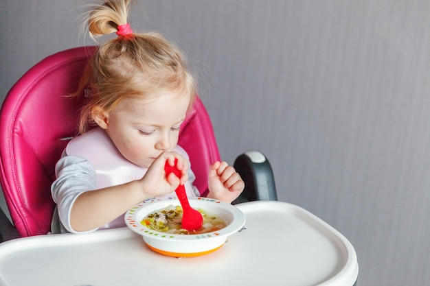 Neonata infantile con la faccia sporca che mangia minestra se stessa con il cucchiaio in seggiolone in cucina a casa