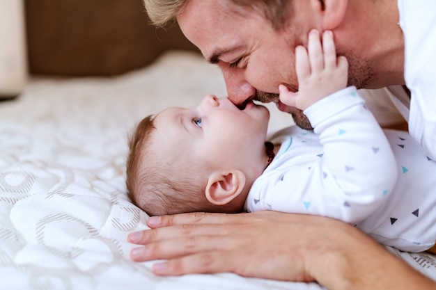 Neonato caucasico adorabile allegro che si trova a letto e che morde il naso di suo padre