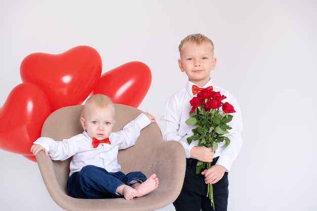 Neonato con palloncini cuore si siede in sedia e fratello ragazzo con bouquet di rose sul muro bianco.