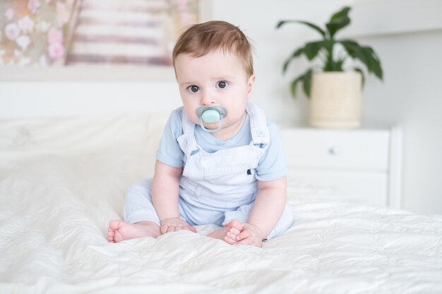 Neonato di 6 mesi con il capezzolo in vestiti blu e seduto sul letto bianco a casa.