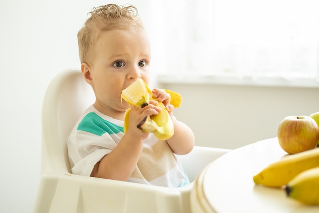 Neonato divertente che si siede alla tavola in sedia del bambino che mangia banana sulla cucina bianca.
