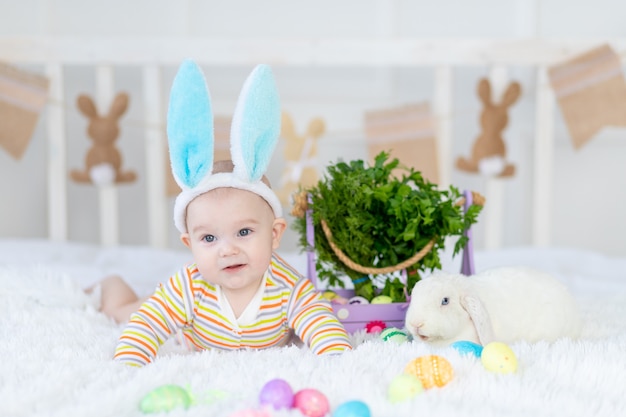 Neonato felice con le orecchie di coniglio sulla sua testa che si trova con un coniglio sul letto con le uova di Pasqua, piccolo bambino sorridente divertente sveglio.