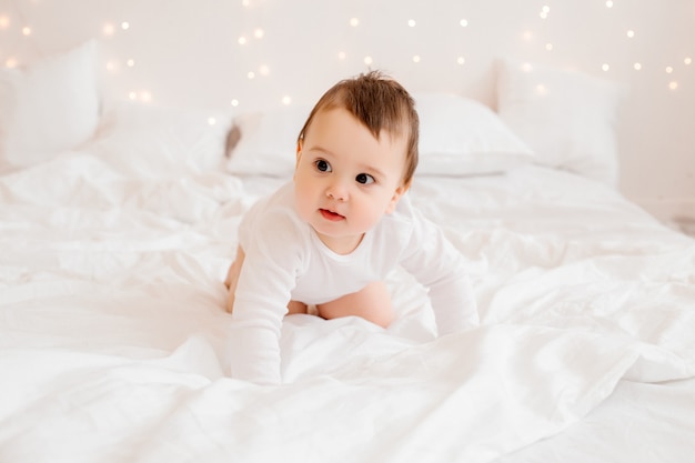 Neonato in buona salute nella seduta sorridente dei vestiti bianchi sulla biancheria da letto bianca a letto