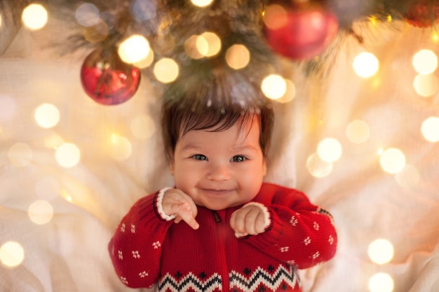 Neonato in un maglione rosso che sorride sotto un albero di Natale con le palle e una ghirlanda a Natale