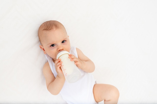 Neonato sdraiato sul letto nella scuola materna sulla schiena e che tiene una bottiglia di latte, alimentando il bambino, concetto di alimenti per l'infanzia