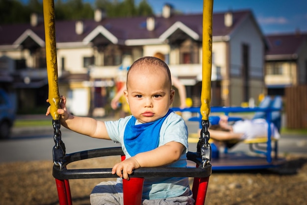 Neonato sorridente felice sveglio sull'oscillazione al parco giochi per bambini all'aperto