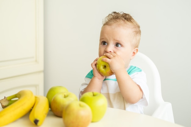 Neonato sveglio che si siede alla tavola in sedia del bambino che mangia mela sulla cucina bianca.