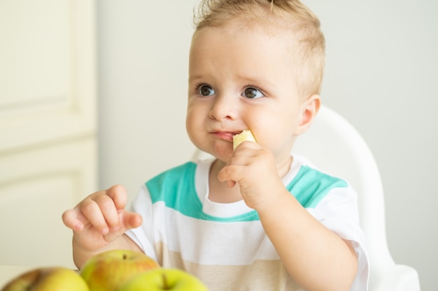 Neonato sveglio che si siede alla tavola nella sedia del bambino che mangia la mela sulla cucina bianca