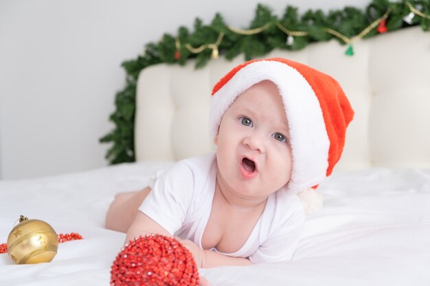 Neonato sveglio che si trova sulla biancheria da letto bianca a casa con il cappello di Natale e le decorazioni di Natale.