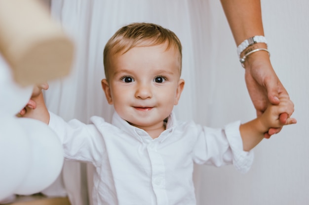 Neonato sveglio in camicia bianca con le mani di sua madre nell'interno luminoso, ritratto alto vicino