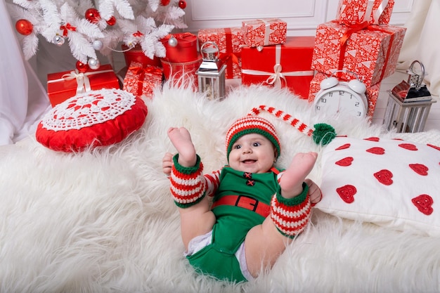 Neonato vestito in costume da gnomo sdraiato su un tappeto di pelliccia bianca tra le decorazioni natalizie