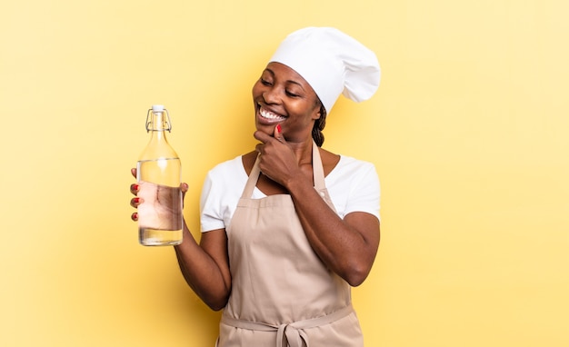 nero chef afro donna sorridente con un'espressione felice e sicura con la mano sul mento, chiedendosi e guardando di lato con in mano una bottiglia d'acqua