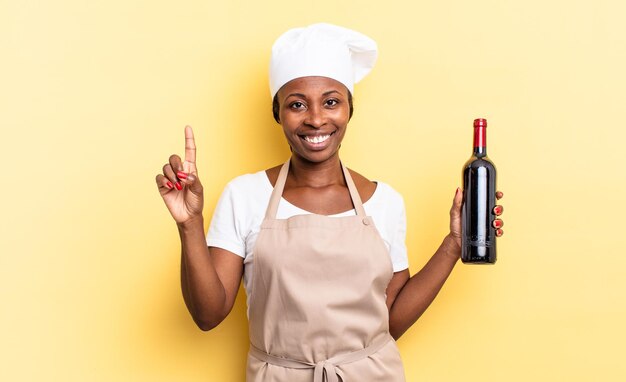 Nero chef afro donna sorridente e dall'aspetto amichevole, mostrando il numero uno o prima con la mano in avanti, conto alla rovescia. concetto di bottiglia di vino