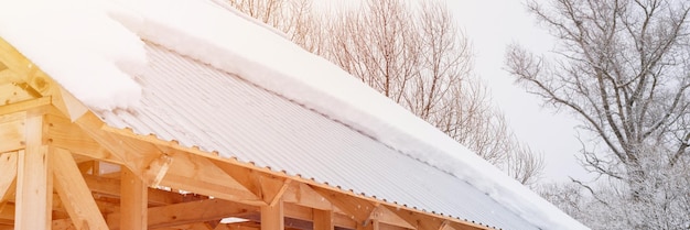 Neve che scivola giù dal tetto edile casa di costruzione con tetto in metallo coperto di neve ghiacciata fresca e fiocchi di neve in una gelida giornata invernale nel sobborgo del villaggio di campagna stagione invernale nevosa banner flare