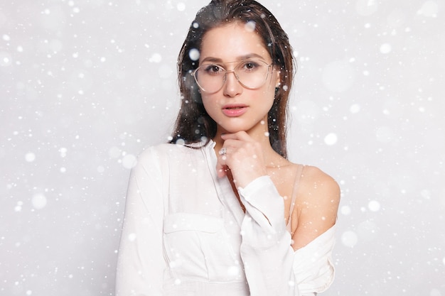 Neve, inverno, natale, persone, concetto di bellezza- Bella ragazza in maglietta bianca bianca, con gli occhiali, parete vuota, ritratto in studio. Sorridente donna bruna con gli occhiali su sfondo di neve