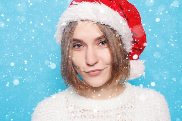 Neve, inverno, natale, persone, concetto di bellezza - cappello natalizio da portare abbastanza biondo su sfondo azzurro di neve. Ritratto di giovane bella ragazza allegra carina che sorride guardando la telecamera