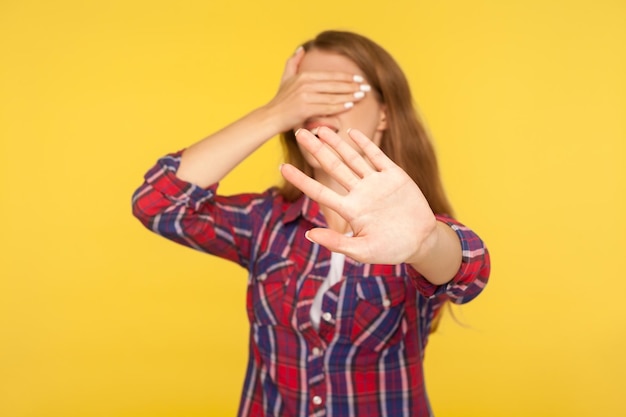 No, non voglio guardare. Ritratto di ragazza in camicia che copre gli occhi con la mano e mostra stop, blocco gesto, sorpresa e paura evitando di guardare. girato in studio isolato su sfondo giallo