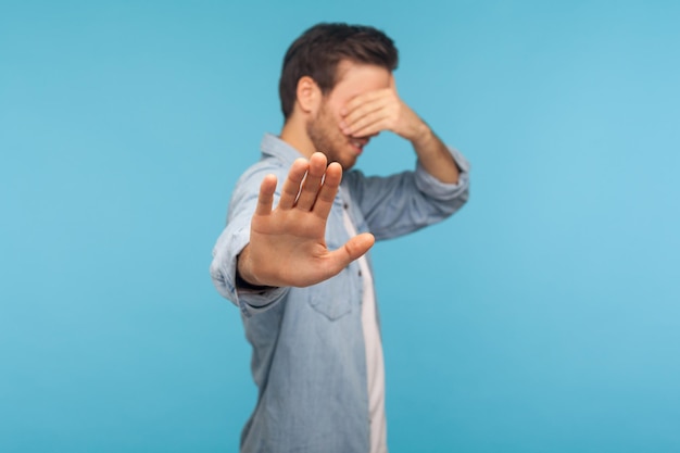 No, non voglio guardare. Ritratto di un uomo timido confuso con una camicia di jeans da lavoratore che copre gli occhi mostrando un gesto di arresto, rifiutandosi di guardare contenuti vergognosi, spaventato di vedere. studio indoor girato, sfondo blu