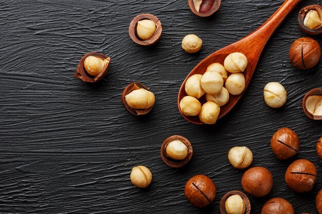 Noce di macadamia sgusciata e noce di macadamia sbucciata su un fondo strutturale nero in un cucchiaio di legno