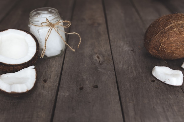 Noci di cocco rotte su fondo di legno grigio con vasetto di olio di cocco extra vergine biologico crudo.