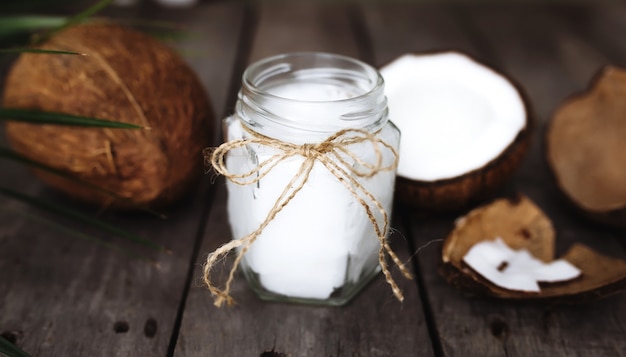 Noci di cocco rotte sulla superficie di legno grigia con vaso di olio di cocco extra vergine biologico crudo e foglia di palma. Polpa di cocco bianca.