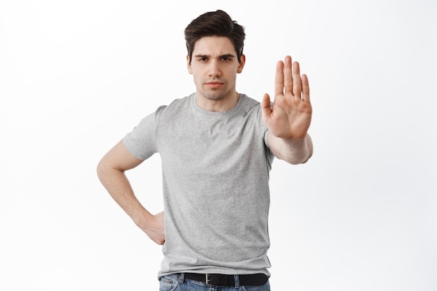 Non avvicinarti, mantieni le distanze. L'uomo sicuro allunga la mano in stop stop, blocca, rifiuta o rifiuta qualcosa con una faccia seria, in piedi su sfondo bianco