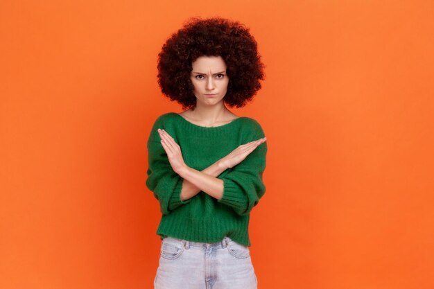 Non c'è modo! Donna seria con acconciatura afro che indossa un maglione verde in piedi con le mani incrociate, che mostra il segno x che significa stop, avviso di problemi. Studio indoor girato isolato su sfondo arancione.