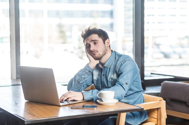 Non lo so! Ritratto di tristezza stanco barbuto giovane libero professionista in camicia di jeans blu sono seduti da soli in un caffè e alla ricerca di un lavoro sul computer portatile sono di cattivo umore senza un buon risultato.