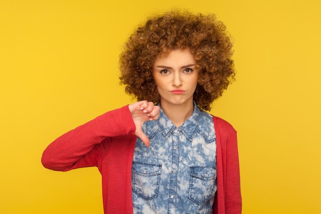 Non mi piace questo Ritratto di donna sconvolta con i capelli ricci in abito casual in piedi con il pollice verso il gesto che esprime disapprovazione criticando il cattivo servizio in studio indoor girato su sfondo giallo