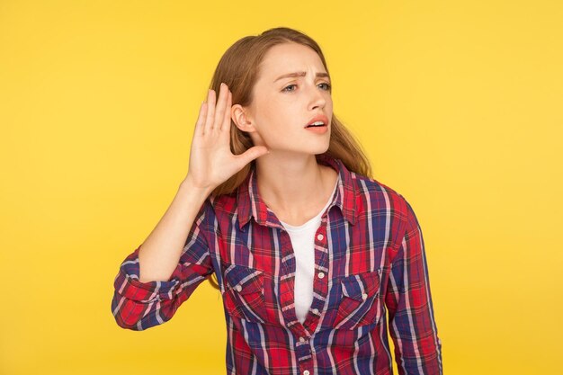 Non riesco a sentirti. Ritratto di ragazza attenta allo zenzero in camicia a scacchi che tiene la mano vicino all'orecchio cercando di ascoltare una conversazione tranquilla, ascoltando pettegolezzi. tiro in studio indoor isolato su sfondo giallo