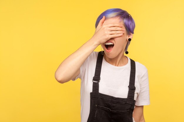 Non voglio guardare Ritratto di una ragazza spaventata e scioccata con i capelli viola in tuta di jeans che copre gli occhi con la mano che esprime insoddisfazione per ciò che vede ignorando il vergognoso contenuto girato in studio