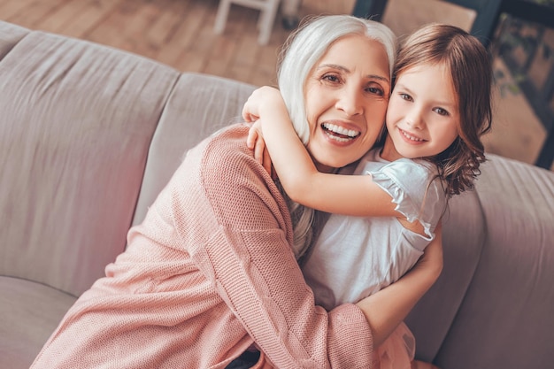 Nonna allegra che abbraccia la nipote seduta sul divano in soggiorno e guardando la telecamera