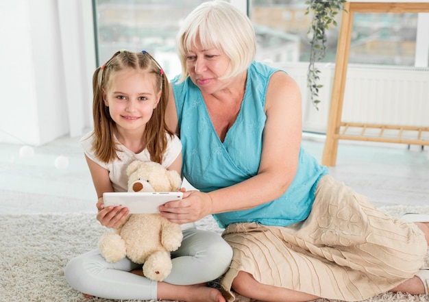 Nonna felice con la piccola nipote utilizzando tablet ubicazione sul pavimento nella stanza dei bambini