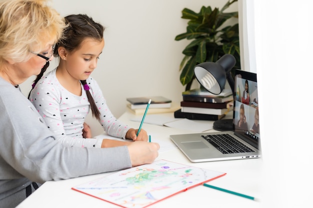 Nonna matura che aiuta il bambino a fare i compiti a casa. Vecchia nonna soddisfatta che aiuta sua nipote a studiare in soggiorno. Bambina che scrive sul taccuino con l'insegnante senior seduto accanto a lei