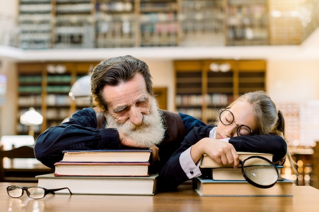 Nonno bell'uomo e nipote bella bambina si addormentò durante la lettura di libri nella vecchia biblioteca antica, seduto al tavolo sopra lo sfondo di scaffali di libri d'epoca