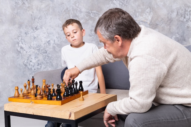 Nonno che gioca vecchi scacchi con il nipote. Il ragazzo e suo nonno sono seduti sul divano in soggiorno e giocano. Un uomo anziano insegna a un bambino a giocare a scacchi