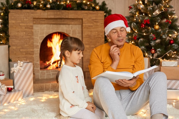 Nonno e nipote che leggono contro il camino e l'albero di Natale nel soggiorno, l'uomo anziano tiene il libro e guarda le pagine con espressione pensierosa, maschio e bambina indossano casualmente.