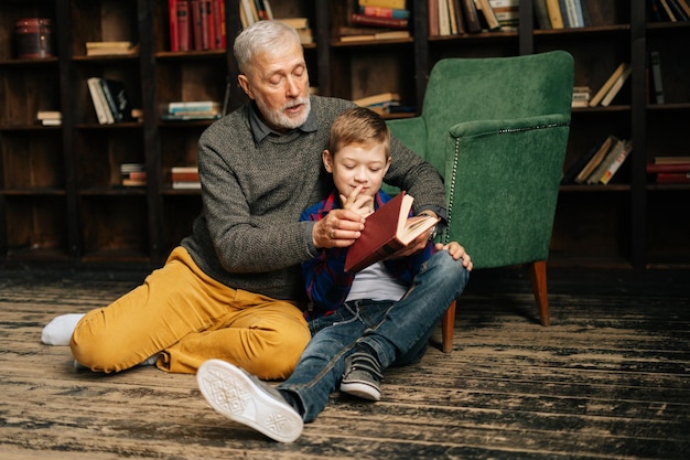 Nonno maturo e nipotino che leggono insieme un libro interessante seduti sul pavimento nella stanza della biblioteca di casa. Libro di lettura del nonno dai capelli grigi barbuto per il nipote.