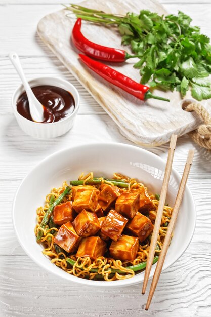 Noodle asiatici dolci e piccanti con tofu croccante al forno hoisin e fagiolini in una ciotola bianca con le bacchette vista verticale dall'alto