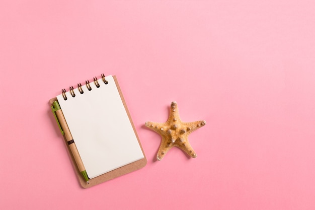 Notebook con conchiglie su sfondo rosa vista dall'alto piatto disteso. Pianificazione delle vacanze estive, del turismo e del concetto di vacanza.