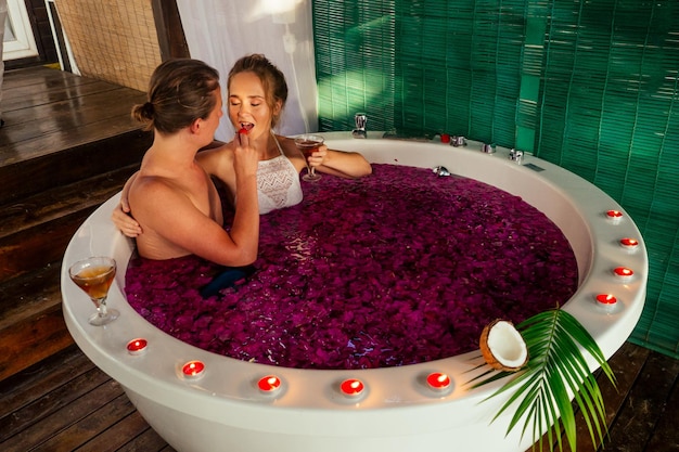 Notte di nozze.bella sposa e sposo bello che si rilassano nella vasca da bagno fiori tropicali al resort di lusso dell'hotel.coppia innamorata di bicchieri tintinnanti eco bali spa.candele,data romantica San Valentino 14 febbraio