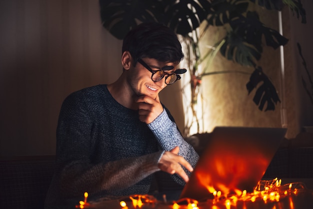 Notte giovane uomo felice con gli occhiali rotondi, guardando nel computer portatile in camera oscura con ghirlande a casa.