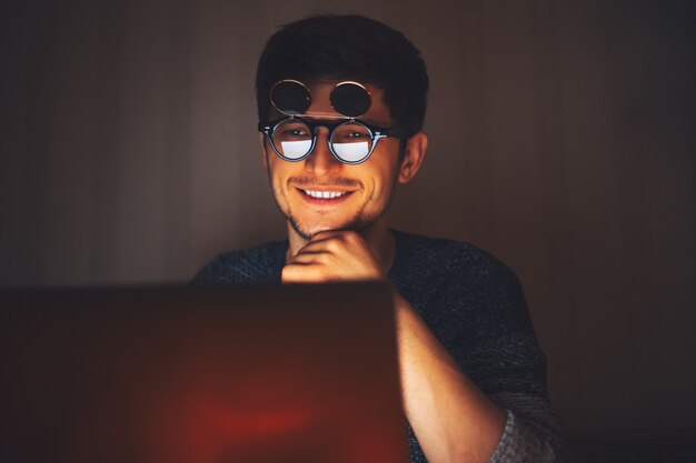 Notte giovane uomo felice con gli occhiali rotondi, guardando nel computer portatile in camera oscura con ghirlande a casa.